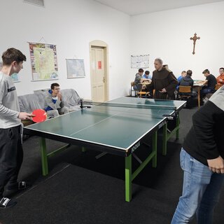 Ein Junge spiel mit einem Franziskanermönch Tischtennis. Ein weiterer Junge sitzt daneben auf einem Sofa und schaut zu. Im Hintergrund spielen 6 weitere Kinder Brettspiele.