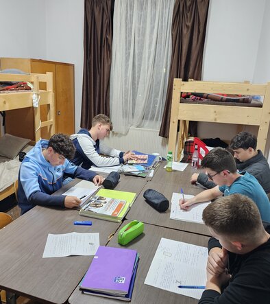 In einem Zimmer mit zwei Stockbetten sitzen 5 Jungen an einem großen Tisch und machen zusammen Hausaufgaben.